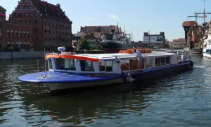 Niebiesko-biały tramwaj wodny "Sonica" płynie po Motławie w Gdańsku, mijając zabytkowe kamienice i inne statki.