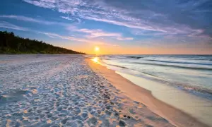 Zachód słońca nad piaszczystą plażą, idealne miejsce dla małych miejscowości nad morzem. Widać ślady na piasku i falujące morze.
