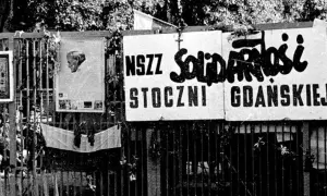 Tablice z napisem "NSZZ Solidarności Stoczni Gdańskiej" wiszą na płocie. Obok portret papieża i inne symbole.