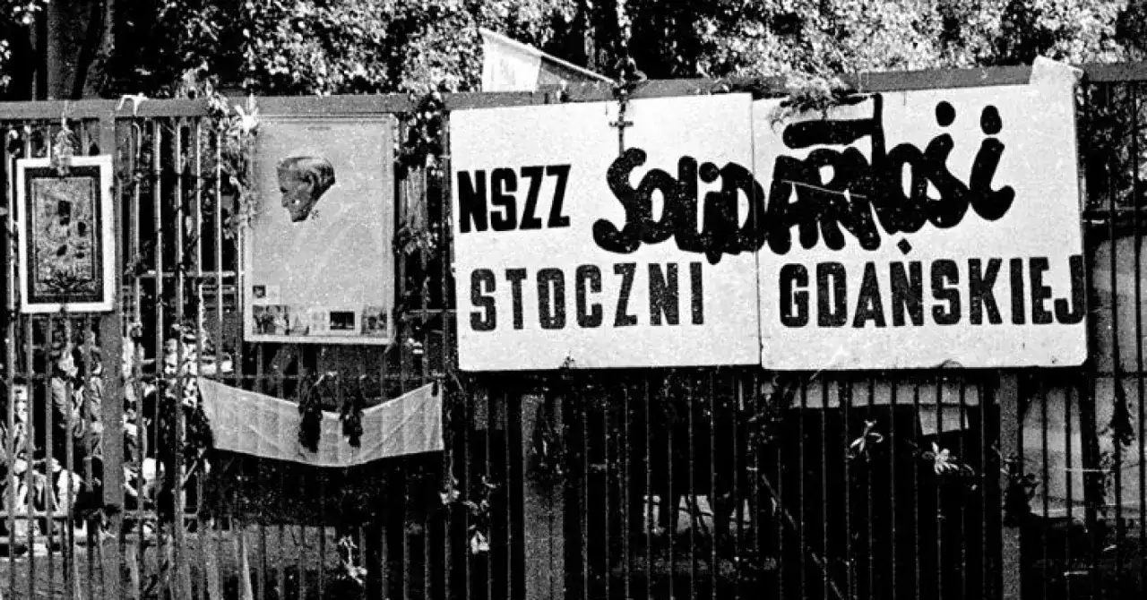 Tablice z napisem "NSZZ Solidarności Stoczni Gdańskiej" wiszą na płocie. Obok portret papieża i inne symbole.
