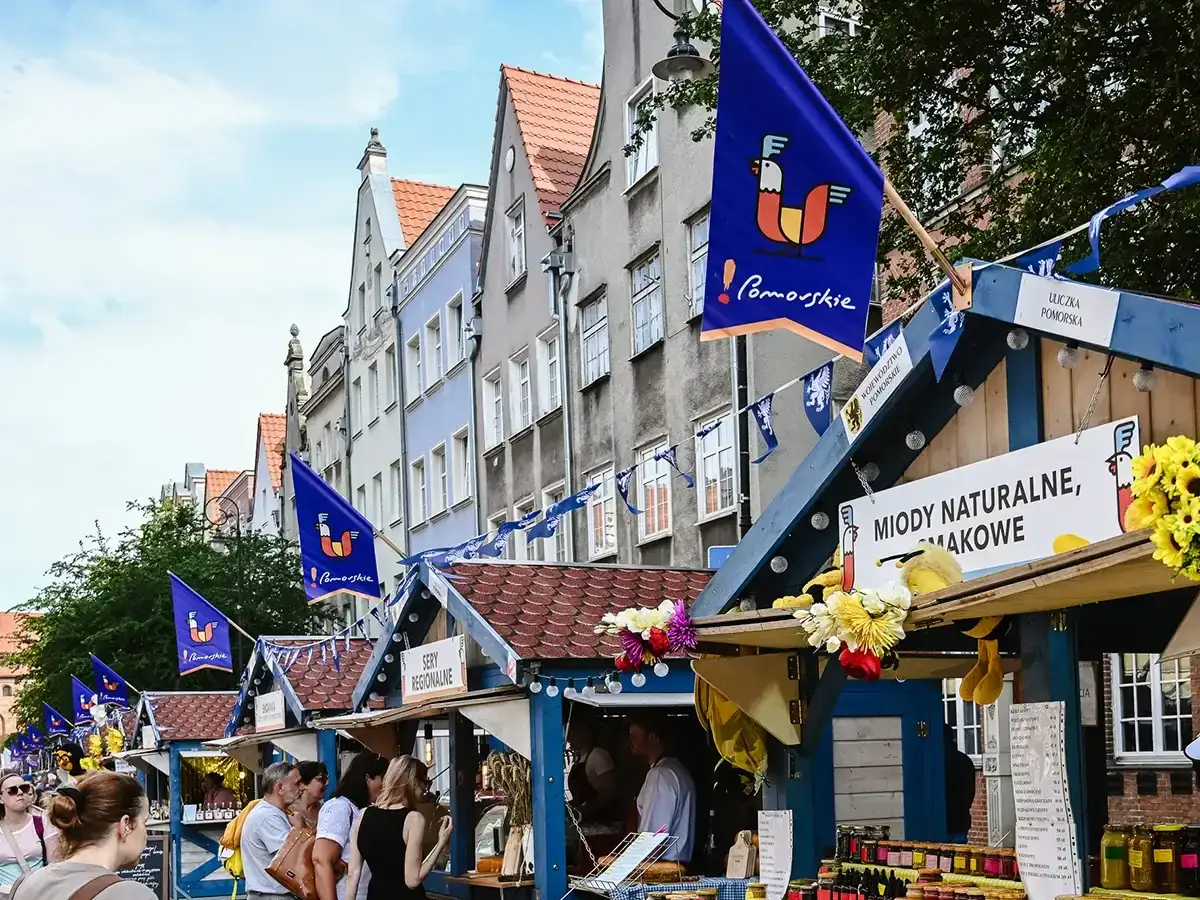Jarmark dominikański w Gdańsku. Kolorowe stragany z miodami i serami, ozdobione flagami z kogutem, na tle zabytkowych kamienic.