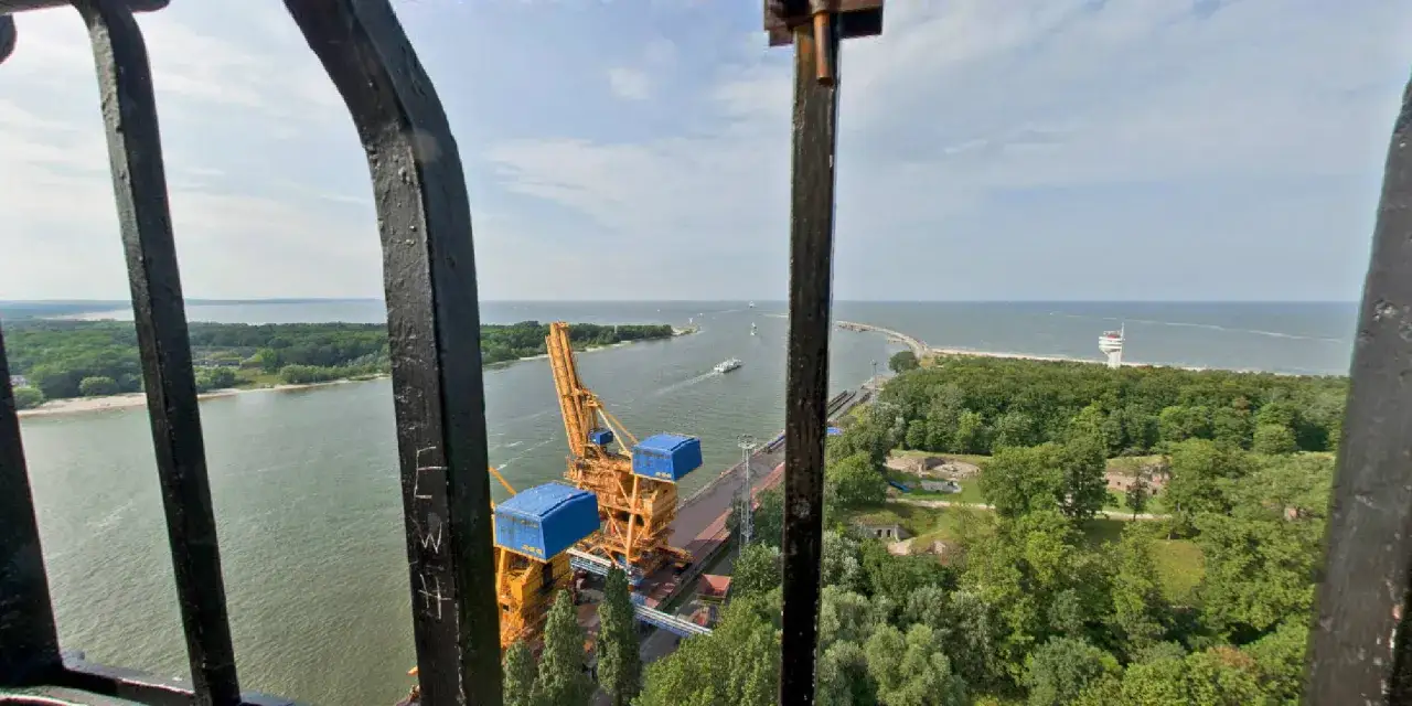 Widok z najwyższej latarni w Polsce na port, morze i zalesione wybrzeże. Widać dźwig portowy i statki.