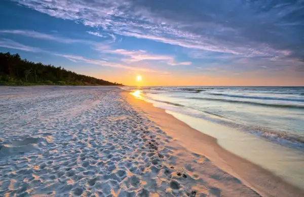 Zachód słońca nad piaszczystą plażą, idealne miejsce dla małych miejscowości nad morzem. Widać ślady na piasku i falujące morze.
